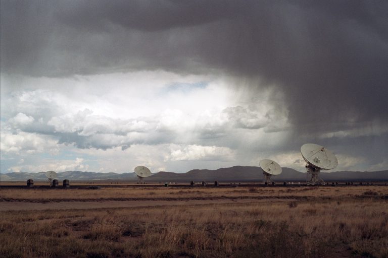 Large radio telescopes stand in a dry, grassy field under a dramatic, cloudy sky with rain in the distance and mountains on the horizon, captured as a striking film photo that highlights nature’s grandeur through professional photography. | Photo by Millworks Film Photography & Prints