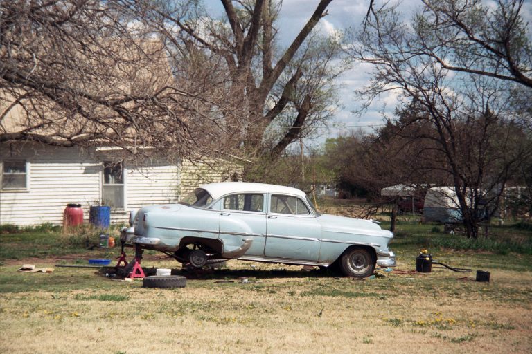 A vintage blue car with missing wheels is propped up on jacks in a grassy yard, surrounded by bare trees, car parts, and a white house in the background—captured in evocative film photography. | Photo by Millworks Film Photography & Prints