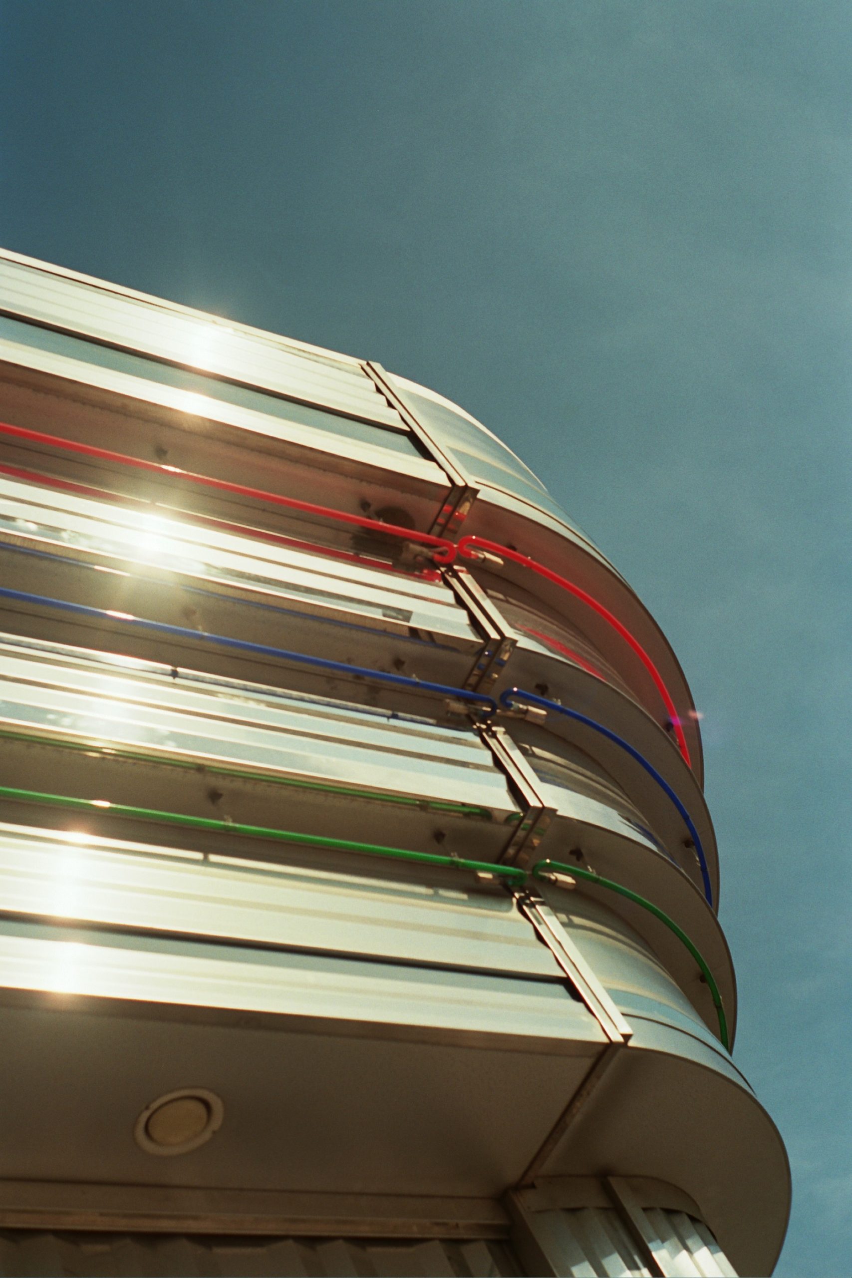 A modern building facade with curved, metallic panels and horizontal blue, red, and green neon lights against a clear blue sky. Sunlight reflects off the shiny surface in this striking example of professional photography. | Photo by Millworks Film Photography & Prints