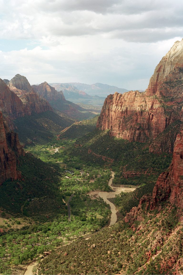 A wide view of a lush green valley with a winding river, surrounded by tall red rock cliffs under a partly cloudy sky in a mountainous landscape, captured through the timeless charm of film photography. | Photo by Millworks Film Photography & Prints