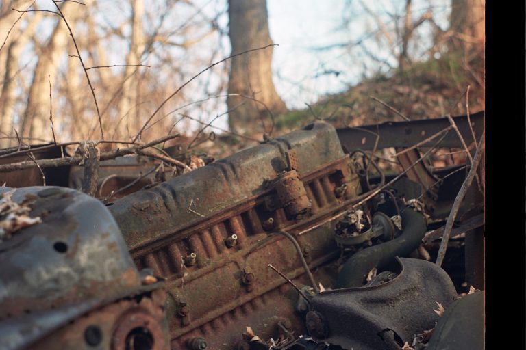 A close-up film photo of a rusty, abandoned car engine lies outdoors among dry branches and leaves, with blurred trees in the background. | Photo by Millworks Film Photography & Prints