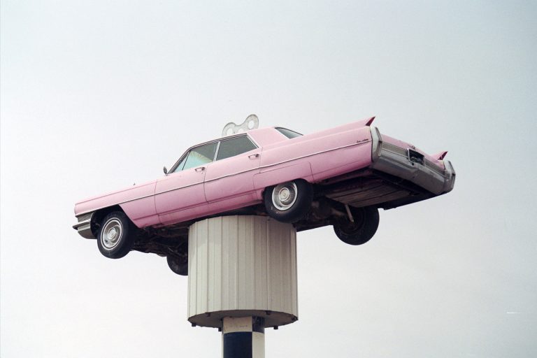 A vintage pink car is mounted high atop a white pole against a pale sky, appearing as though it is floating above the ground—a perfect subject for film photography. | Photo by Millworks Film Photography & Prints