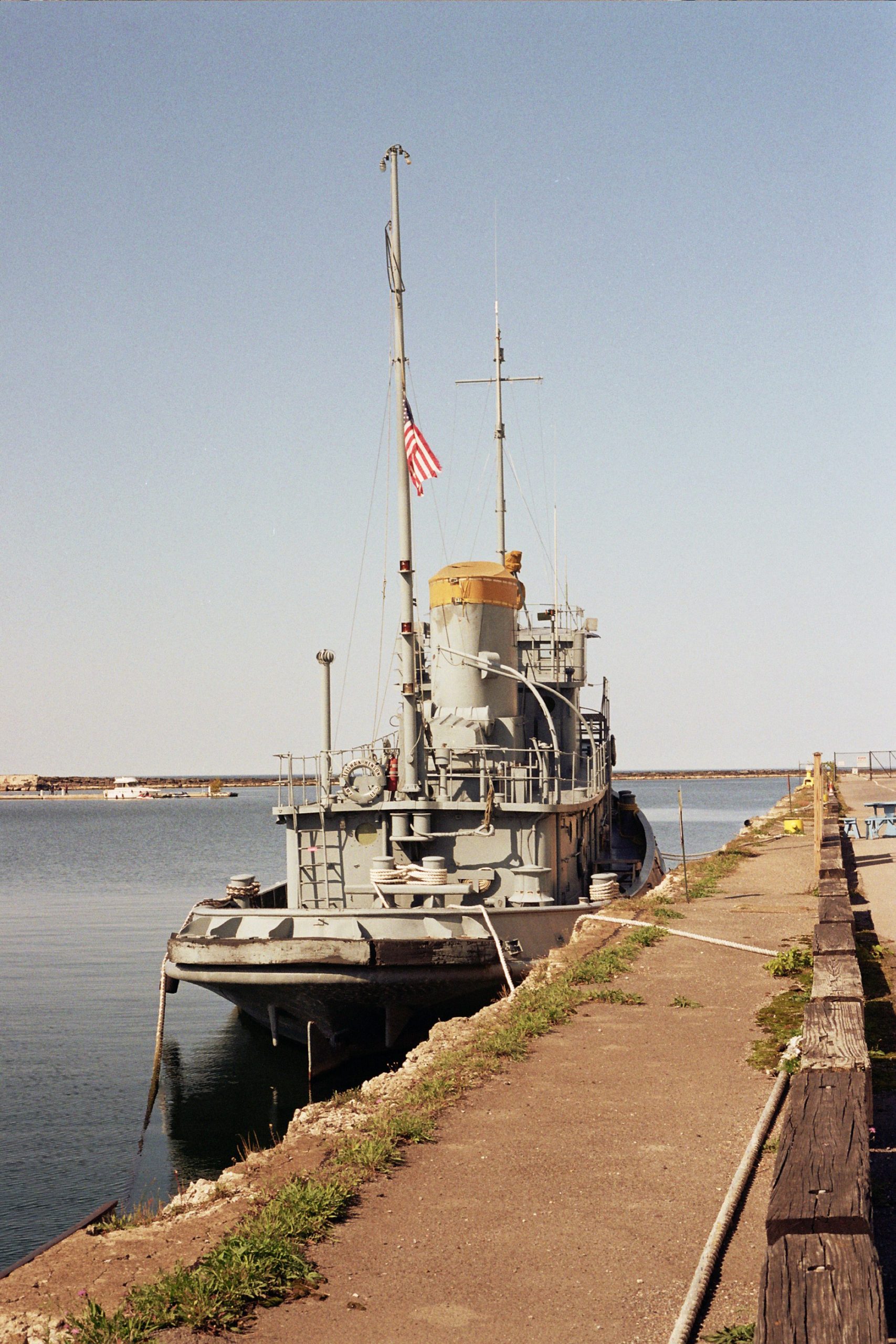 A gray military ship with an American flag is docked at a concrete pier on a calm, sunny day. Captured with film photography, the clear blue sky and water bring a timeless quality to this peaceful scene. | Photo by Millworks Film Photography & Prints
