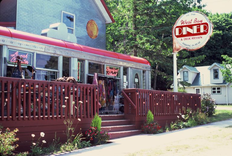 A vintage diner with a red railing and outdoor deck displays racks of clothes. Captured in the style of film photography, a large round sign reads West Bay Diner & Delicatessen, with trees and houses visible in the background. | Photo by Millworks Film Photography & Prints