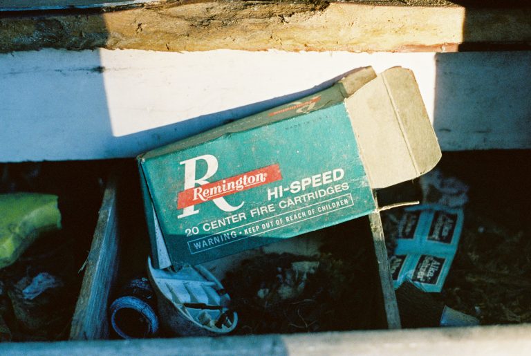 An old, partially crushed Remington Hi-Speed cartridge box lies in a drawer filled with scattered debris and miscellaneous items, sunlight casting shadows across the scene like a timeless film photo. | Photo by Millworks Film Photography & Prints