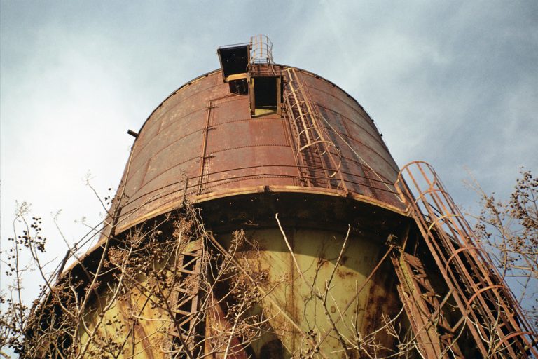 A low-angle view of a large, rusted industrial water tower with metal ladders and platforms, surrounded by dry branches against a cloudy sky, captured in the timeless style of film photography. | Photo by Millworks Film Photography & Prints