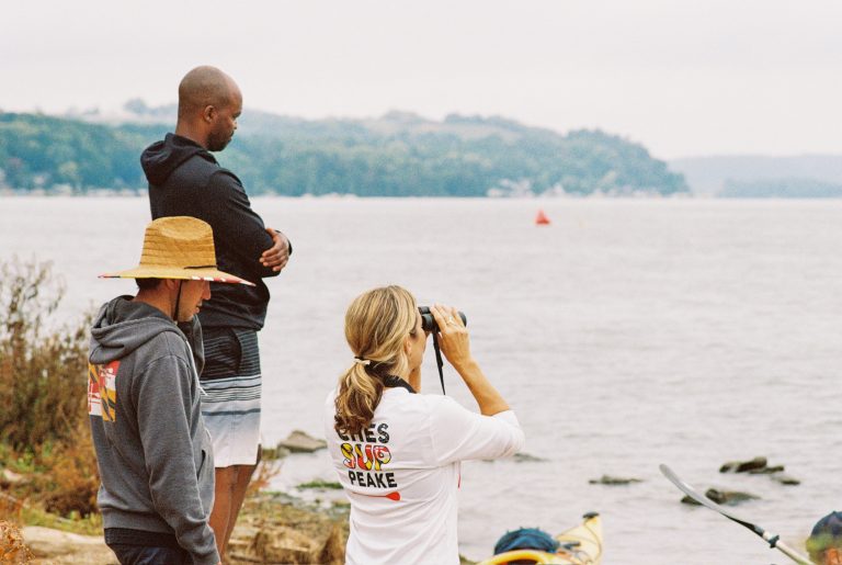 Three people stand on a rocky shoreline by a large body of water. One woman uses binoculars, a man stands with arms crossed, and another person wears a wide-brim hat. Kayaks rest at the waters edge in this striking film photography scene. | Photo by Millworks Film Photography & Prints