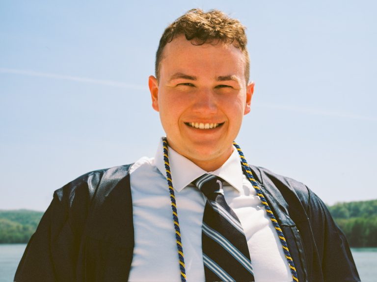 A smiling young man in a graduation gown, honor cords, and a striped tie stands outdoors by a lake and trees under a clear sky, captured in classic film photography style. | Photo by Millworks Film Photography & Prints
