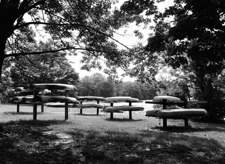 Several canoes are stacked on wooden racks under large leafy trees by a calm lake. Captured with film photography, the scene is serene, with dappled sunlight and abundant greenery enhancing its timeless charm. | Photo by Millworks Film Photography & Prints