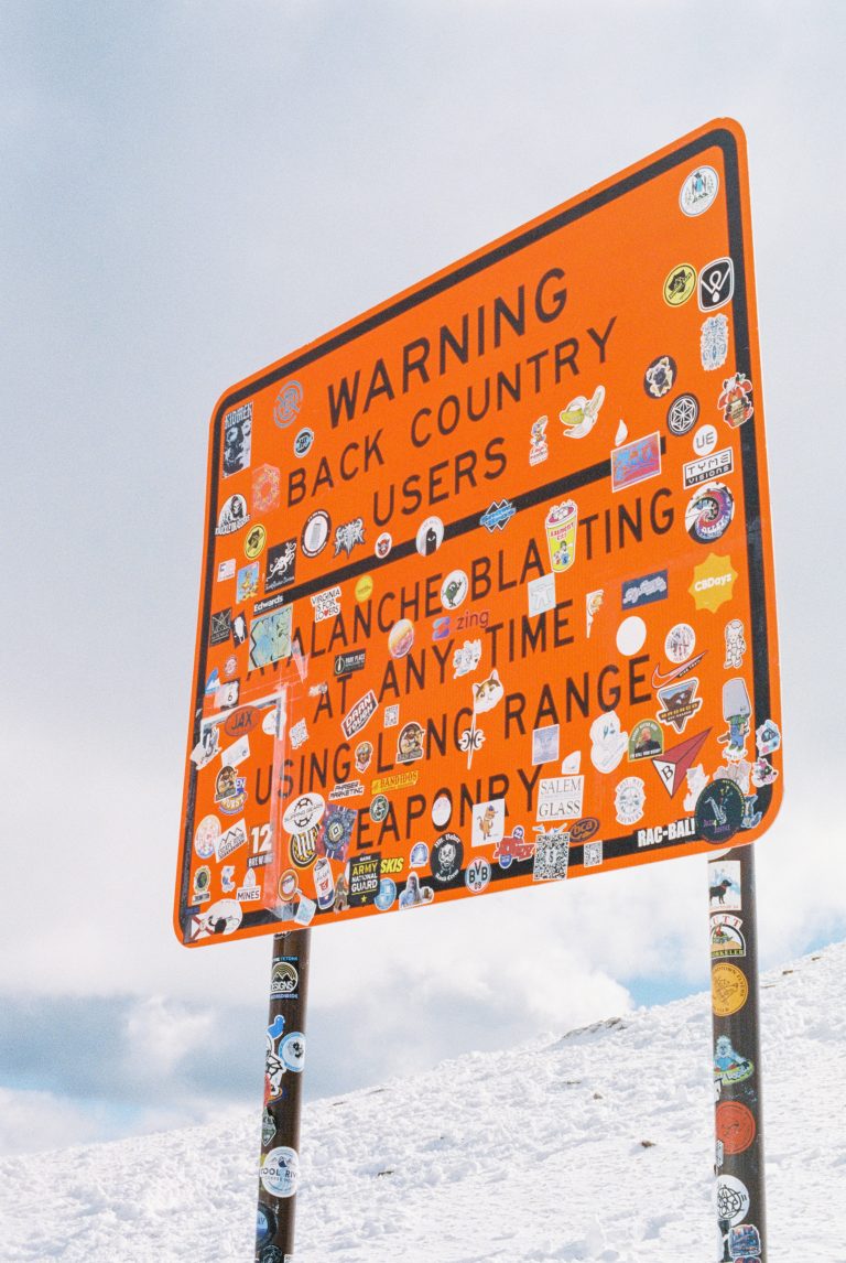 Large orange warning sign in snowy landscape, covered with colorful stickers. Captured in crisp film photography, the sign warns backcountry users about avalanche blasting and weapon range under a blue sky with clouds. | Photo by Millworks Film Photography & Prints