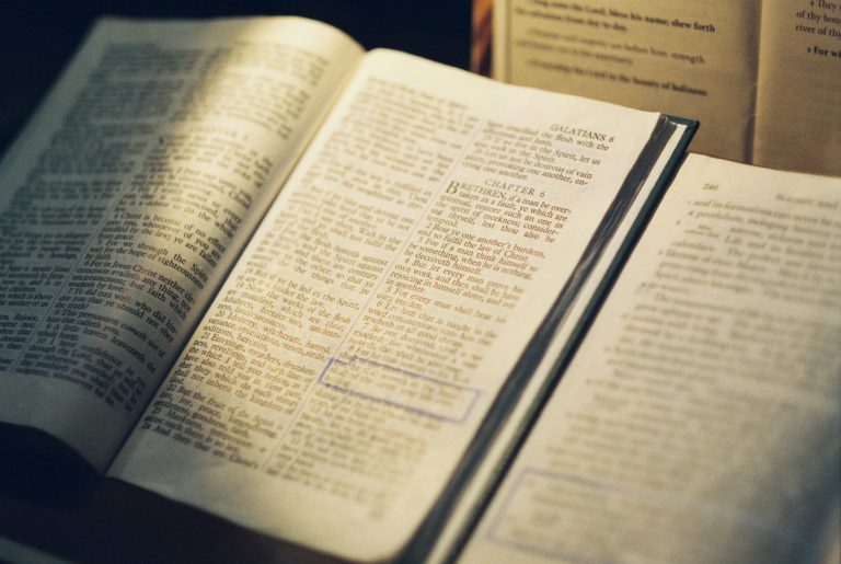 An open Bible with highlighted passages and visible chapter headings rests on a table beside another open book. Warm lighting illuminates the pages, capturing the scene in the timeless style of film photography. | Photo by Millworks Film Photography & Prints