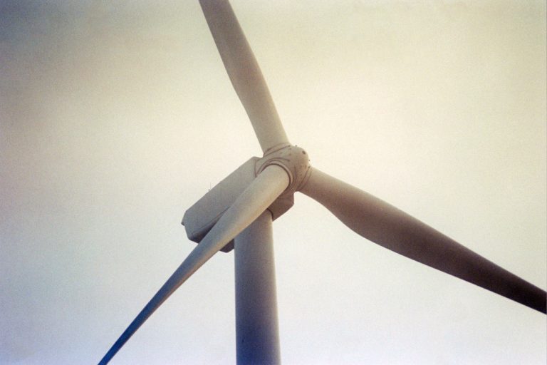Close-up view of a wind turbine with three blades against a light, slightly hazy sky. Captured from a low angle, the image showcases the size and structure of the turbine in classic film photography style. | Photo by Millworks Film Photography & Prints