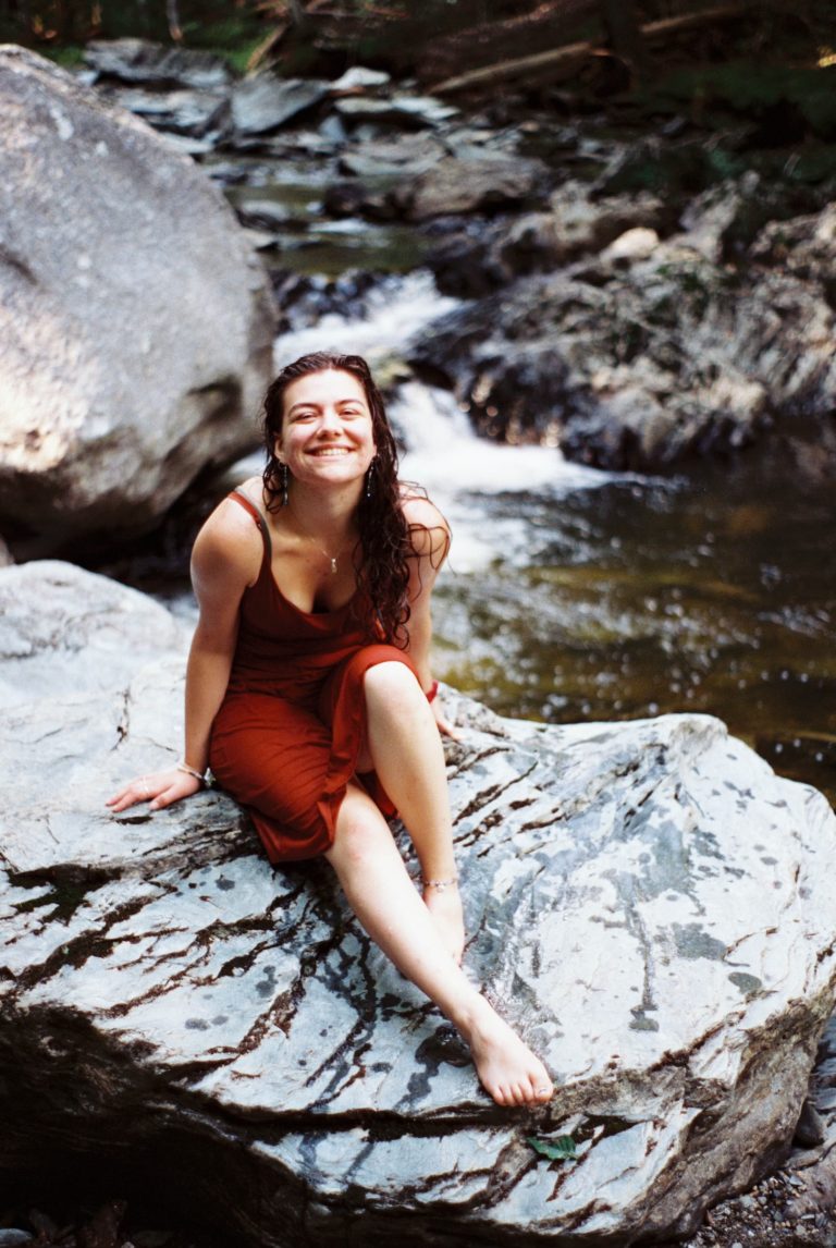 A woman in a red dress sits barefoot on a large rock by a flowing creek, smiling brightly at the camera. Lush greenery and rocks surround the water in this captivating film photo. | Photo by Millworks Film Photography & Prints