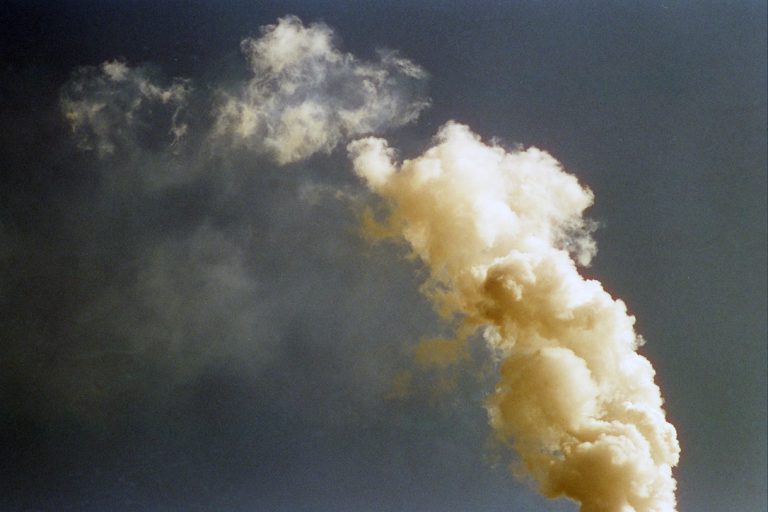 A large plume of thick, yellowish smoke rises against a dark sky, gradually dispersing as it moves upward—a scene reminiscent of classic film photography. | Photo by Millworks Film Photography & Prints