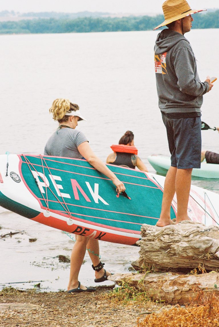A woman carries a teal and red paddleboard labeled PEAK toward the water, captured in striking film photography, while a man in a straw hat stands on a log nearby. Other people paddle in the background on a calm, overcast day. | Photo by Millworks Film Photography & Prints