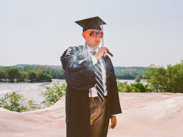 A person in a graduation cap and gown, wearing bright sunglasses, stands outdoors by water and greenery, holding and smoking a cigar—sunlit smoke curls poetically in this vibrant film photography scene. | Photo by Millworks Film Photography & Prints
