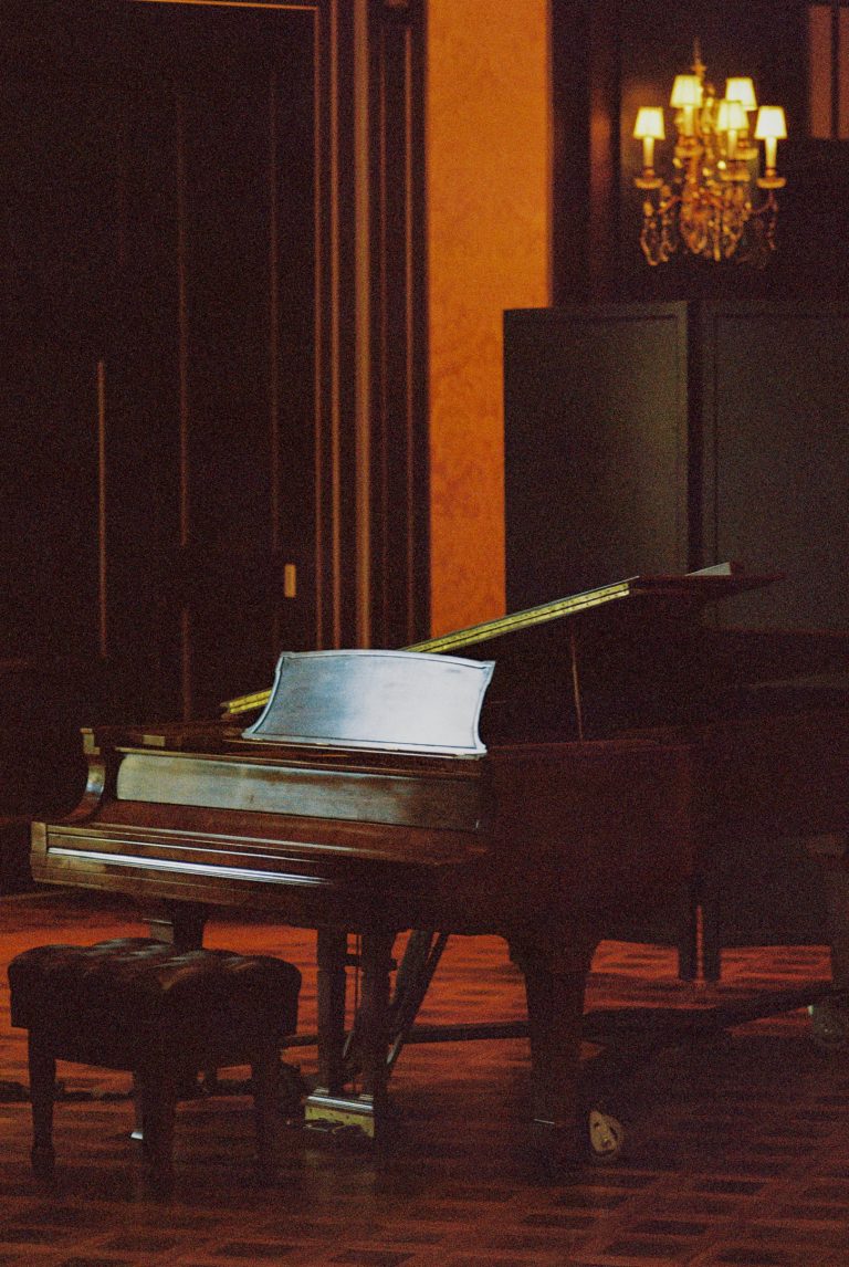 A grand piano and a piano bench sit on a polished wooden floor in a dimly lit room with dark walls, captured in the warm glow of a chandelier—an inviting scene reminiscent of classic film photography. | Photo by Millworks Film Photography & Prints