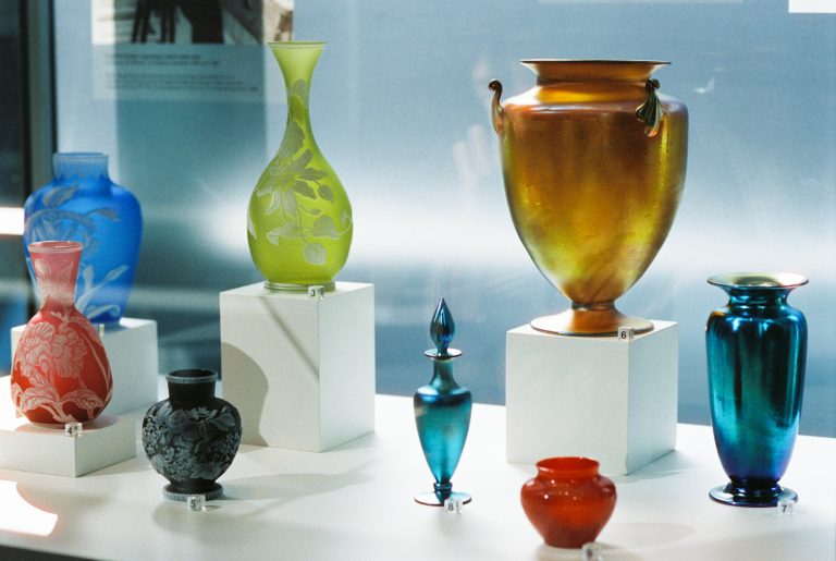 A display of colorful glass vases and vessels in various shapes and sizes, arranged on white pedestals in a well-lit exhibition setting. Captured through professional photography, the vibrant blues, greens, oranges, reds, and golds stand out brilliantly. | Photo by Millworks Film Photography & Prints