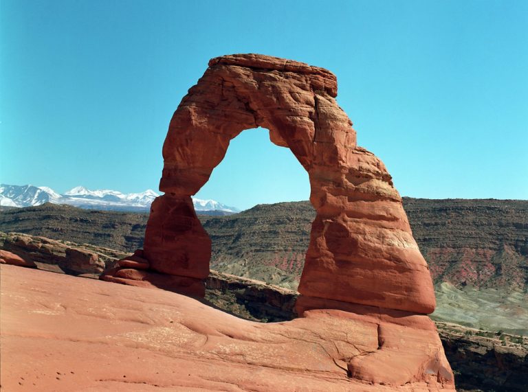 Red sandstone arch, known as Delicate Arch, stands on a rocky plateau under a clear blue sky, captured in stunning professional photography with distant snow-capped mountains and layered desert hills in the background. | Photo by Millworks Film Photography & Prints