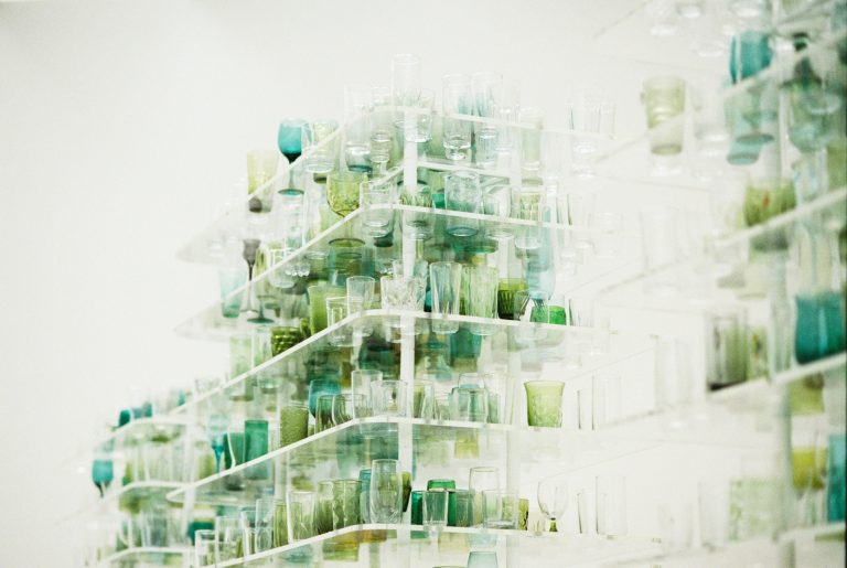 Shelves filled with various transparent and greenish glass cups and tumblers are stacked closely together in an abstract, artistic arrangement against a white background, capturing the scene with a film photography aesthetic. | Photo by Millworks Film Photography & Prints