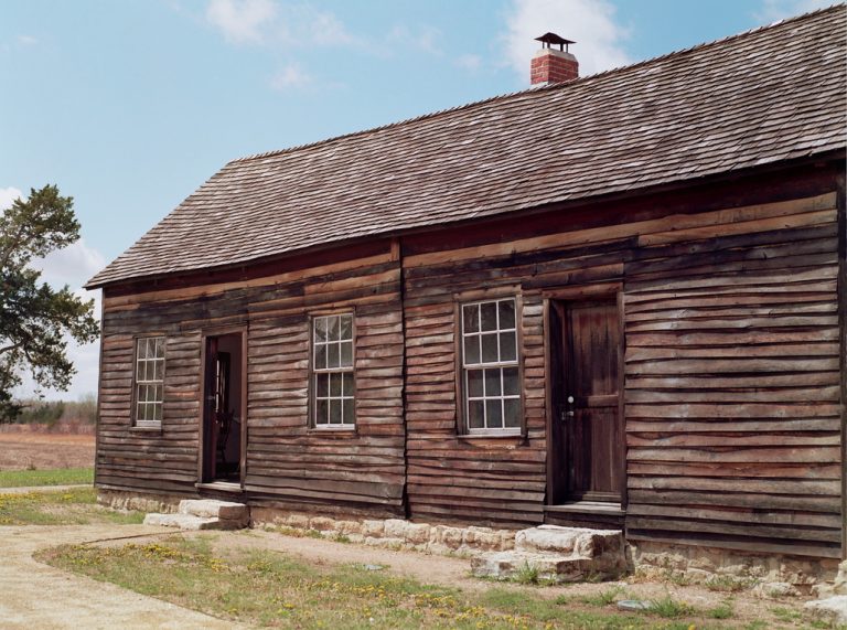 A rustic wooden house with a shingled roof, two windows, and two doors stands on a stone foundation in a rural landscape under a partly cloudy sky—captured in the timeless style of film photography. | Photo by Millworks Film Photography & Prints