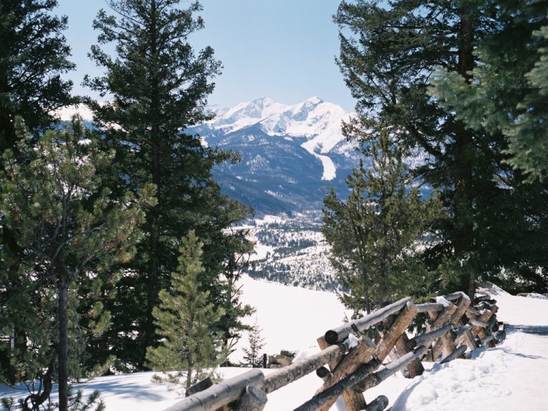 A rustic wooden fence lines a snow-covered path among pine trees, with distant snow-capped mountains under a clear blue sky—captured in the timeless charm of film photography. | Photo by Millworks Film Photography & Prints
