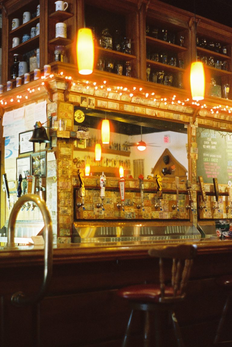 A cozy bar with hanging amber lights, a row of beer taps, and shelves filled with glasses and mugs, captured in the soft glow of film photography. A high wooden chair sits in front of the counter, adding to the inviting atmosphere. | Photo by Millworks Film Photography & Prints