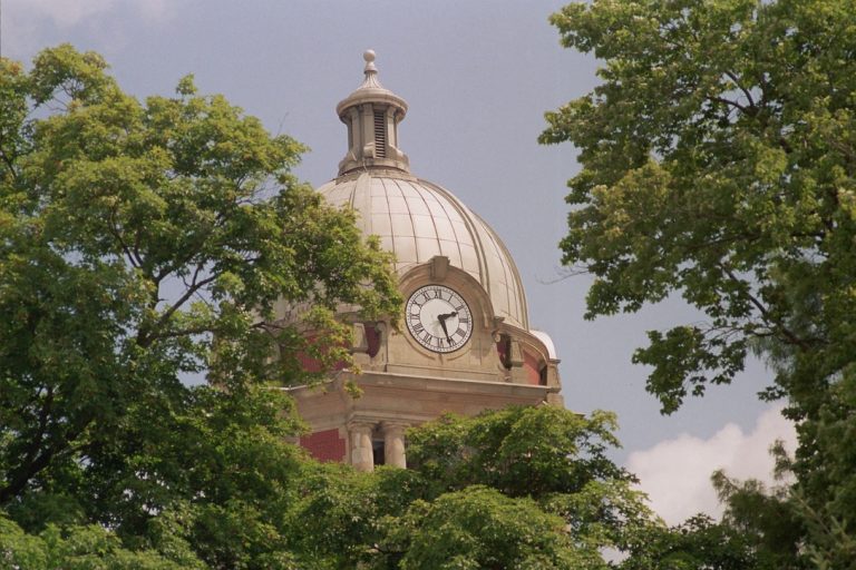 A domed clock tower, partially obscured by leafy green trees, stands against a blue sky with clouds in this striking film photography shot. The clock shows the time as 4:35. | Photo by Millworks Film Photography & Prints