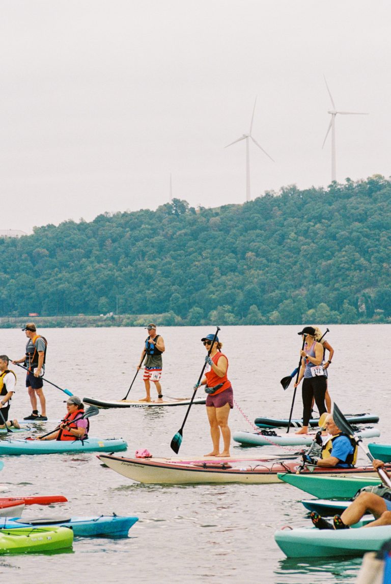 A group of people paddleboarding and kayaking on a lake, wearing life jackets and helmets, is captured in this professional photography shot with forested hills and wind turbines in the background under a cloudy sky. | Photo by Millworks Film Photography & Prints