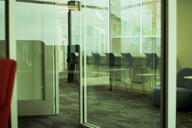 A modern, empty office space with glass walls, high-backed chairs, small round tables, and large windows letting in natural light—captured in the soft tones of film photography. The atmosphere appears quiet and open. | Photo by Millworks Film Photography & Prints