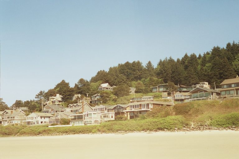 Houses line a hillside covered with trees, overlooking a sandy beach under a clear blue sky, captured in the timeless style of film photography. | Photo by Millworks Film Photography & Prints