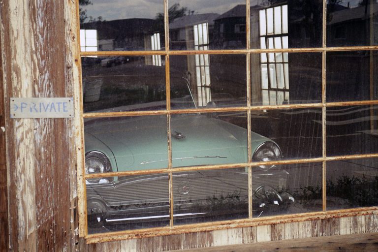 A vintage green car is visible through the large, grid-paned window of a weathered wooden garage. Captured as a film photo, the scene features a PRIVATE sign posted on the exterior wall beside the window. | Photo by Millworks Film Photography & Prints