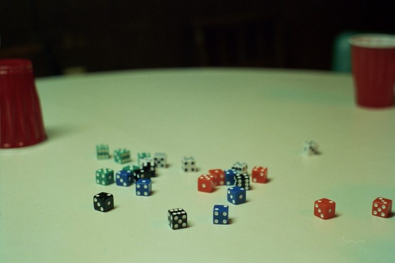 Scattered dice of various colors are spread across a round off-white table. Captured in the style of film photography, two upside-down red plastic cups rest nearby, with some chairs visible in the background. | Photo by Millworks Film Photography & Prints