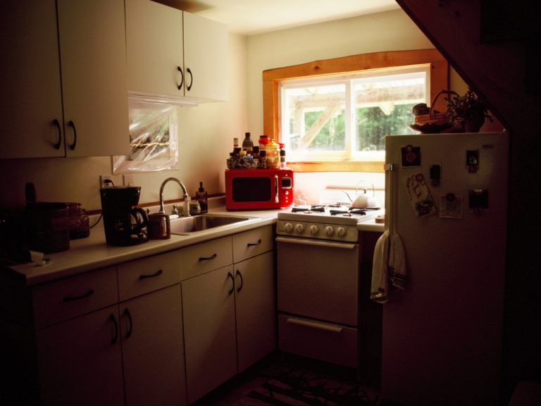 A small kitchen with white cabinets, a sink, stove, red microwave, and refrigerator. Sunlight streams through a window above the sink, illuminating the countertops in a way that evokes classic film photography charm. | Photo by Millworks Film Photography & Prints