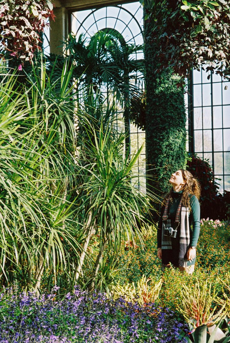 A woman stands among lush green plants and colorful flowers in a sunlit greenhouse, gazing upward with sunlight streaming through large arched windows behind her in this stunning film photography scene. | Photo by Millworks Film Photography & Prints