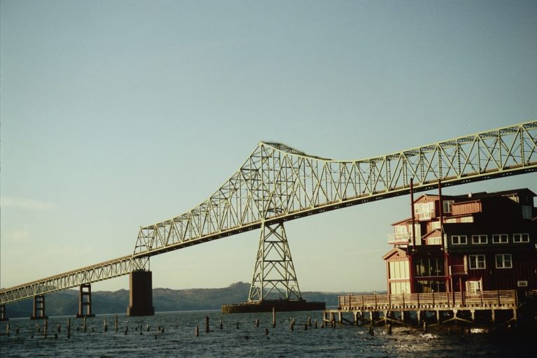 A large truss bridge spans a wide river, with part of the bridge passing above a red building on the water’s edge, under a clear sky. Wooden pilings are visible below in this stunning example of professional photography. | Photo by Millworks Film Photography & Prints