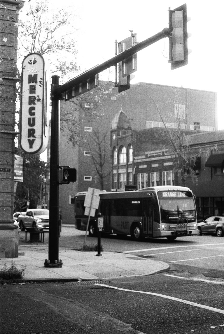 A black-and-white film photo of a city street corner shows a bus labeled Orange Line turning at an intersection near a vertical Mercury sign and traffic lights. Buildings and trees line the background, capturing classic film photography style. | Photo by Millworks Film Photography & Prints