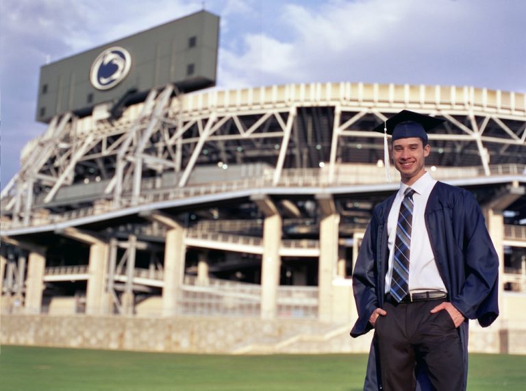 A young man in a graduation cap and gown smiles while standing on grass in front of a large football stadium with a Penn State logo on the scoreboard, captured in stunning professional photography. | Photo by Millworks Film Photography & Prints