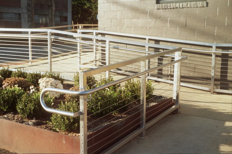 A metal handrail ramp with horizontal cables is next to a raised garden bed with green shrubs and white flowers, beside a concrete building and sidewalk, captured in the soft tones of film photography. | Photo by Millworks Film Photography & Prints