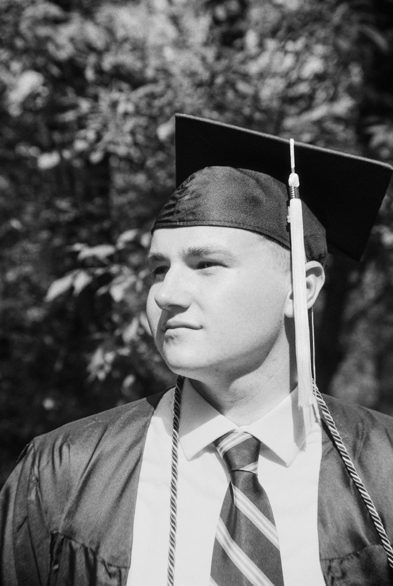 A young person in a graduation cap and gown looks to the side, standing outdoors with trees in the background. Wearing a striped tie and graduation cord, they are captured in this black and white film photo. | Photo by Millworks Film Photography & Prints