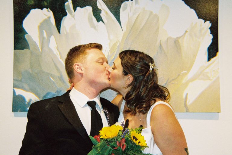 A bride and groom share a kiss in front of a large white flower painting. Captured on film photography, the groom wears a black suit and tie while the bride, with her hair pinned back, holds a bouquet of sunflowers in her white dress. | Photo by Millworks Film Photography & Prints