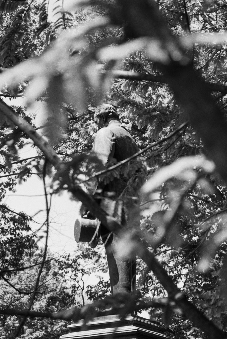 A black and white film photo of a statue of a man holding a hat, partially obscured by tree branches and leaves in the foreground. The background is filled with trees and dappled sunlight, capturing the classic charm of film photography. | Photo by Millworks Film Photography & Prints