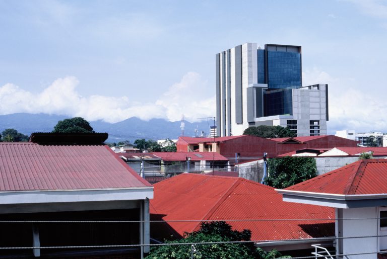 A cityscape with red-roofed buildings in the foreground and a tall, modern glass and concrete building in the background, set against distant mountains and a partly cloudy sky, captured with the classic look of film photography. | Photo by Millworks Film Photography & Prints