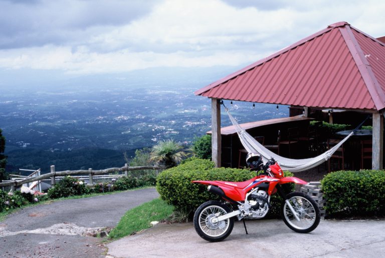 A red motorcycle is parked near a gazebo with a hammock, overlooking a scenic valley and mountains under a cloudy sky—captured in the timeless style of film photography. | Photo by Millworks Film Photography & Prints