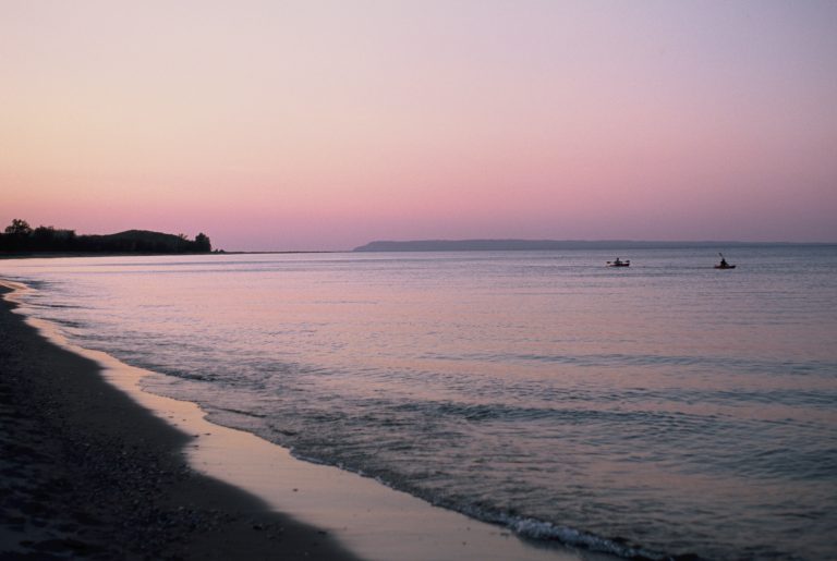A calm beach at sunset with soft pink and purple hues in the sky, captured in a stunning film photo. Gentle waves touch the shore, two small boats float on tranquil water, and low hills are visible on the horizon. | Photo by Millworks Film Photography & Prints