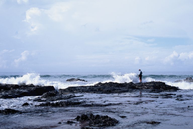 A person stands on a rocky shore as waves crash around them under a cloudy sky, facing the ocean. Captured in the timeless style of film photography, the serene and expansive scene highlights the beauty of water and rocks in the foreground. | Photo by Millworks Film Photography & Prints