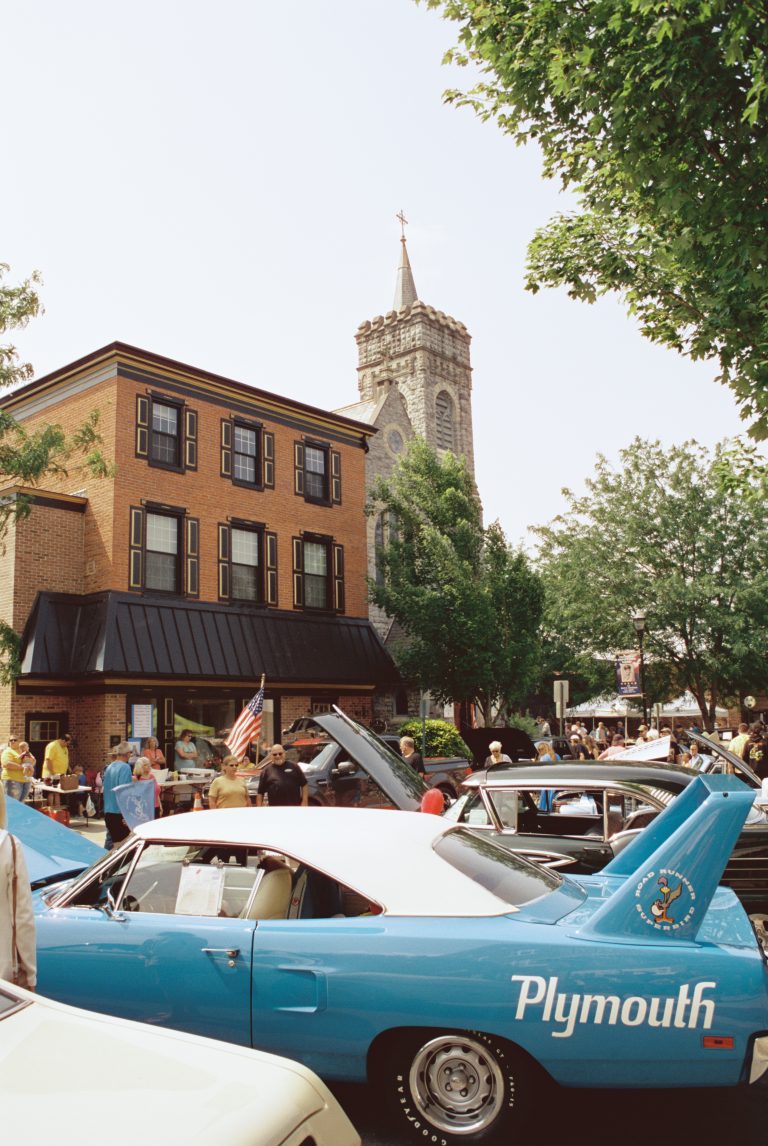 People gather at a car show featuring classic vehicles, including a blue Plymouth with white accents. Captured with film photography, the scene includes a brick building and a church steeple shaded by green trees. | Photo by Millworks Film Photography & Prints