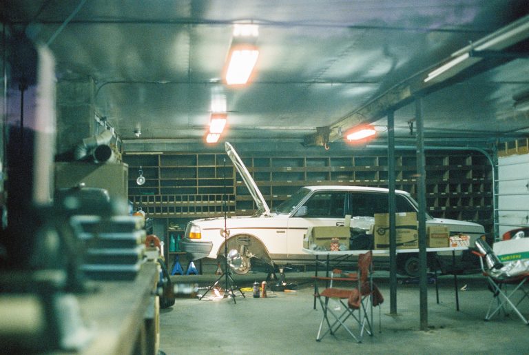 A vintage white car with its hood open is parked in a dimly lit garage. Tools and equipment are scattered around, and folding chairs sit nearby. Fluorescent lights illuminate the workspace in this atmospheric film photo. | Photo by Millworks Film Photography & Prints