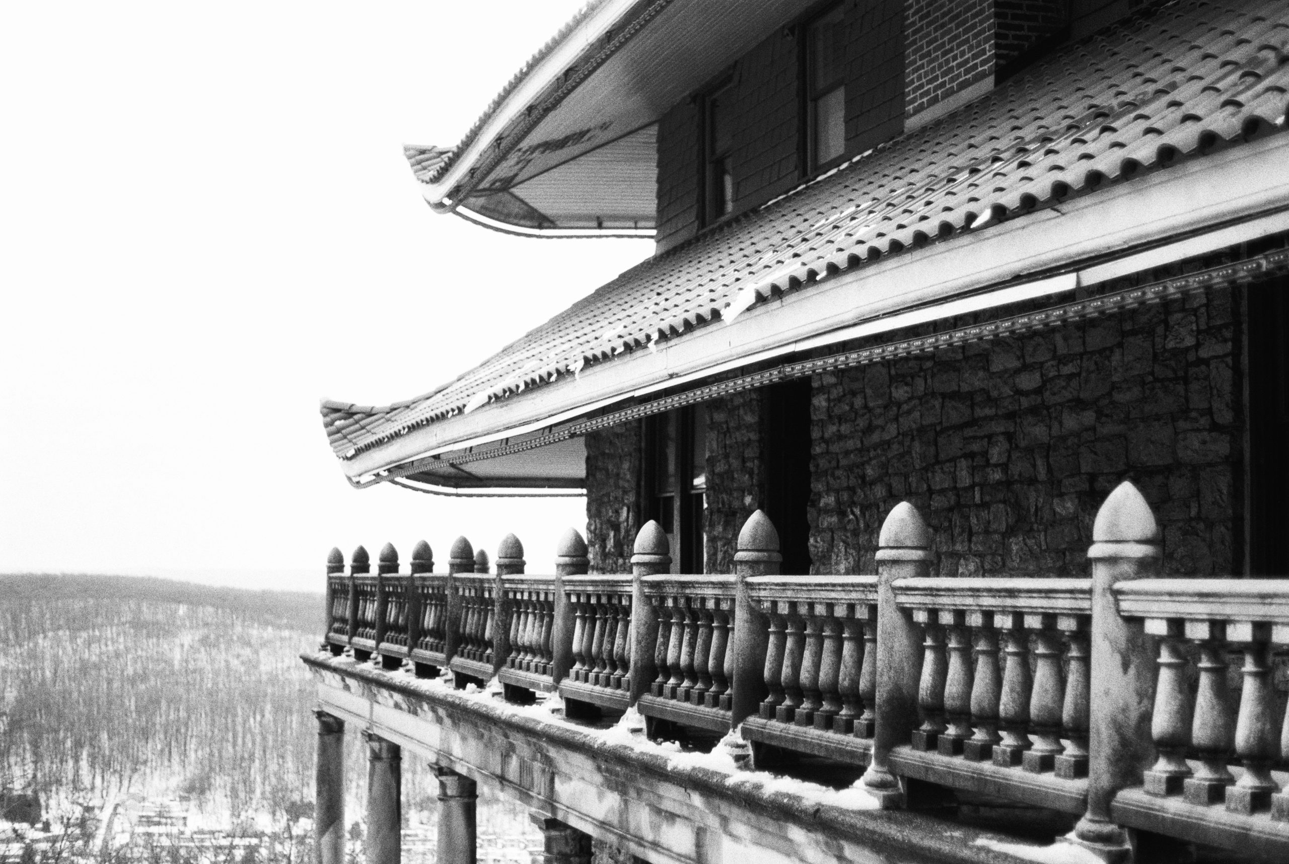 A black and white film photo of a building with Asian-inspired curved rooflines, stone walls, and a decorative balustrade balcony overlooking a snowy, tree-covered landscape. | Photo by Millworks Film Photography & Prints