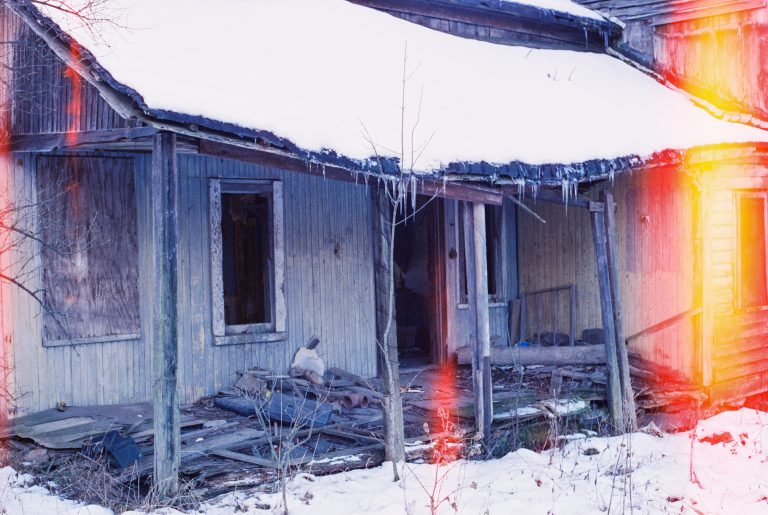 An old, abandoned wooden house with a sagging, snow-covered porch roof, broken windows, and scattered debris. Icicles hang from the roof as streaks of light illuminate the scene, evoking the nostalgic charm of film photography. Snow covers the ground. | Photo by Millworks Film Photography & Prints